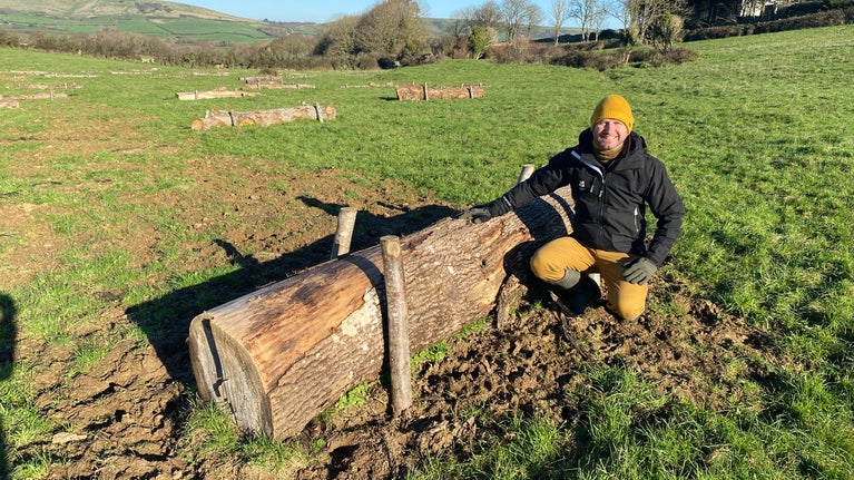Ranger demonstrating surface roughening - he is crouching beside a log that has been laid in a field to slow down the flow of water. Other logs are laid out behind him.